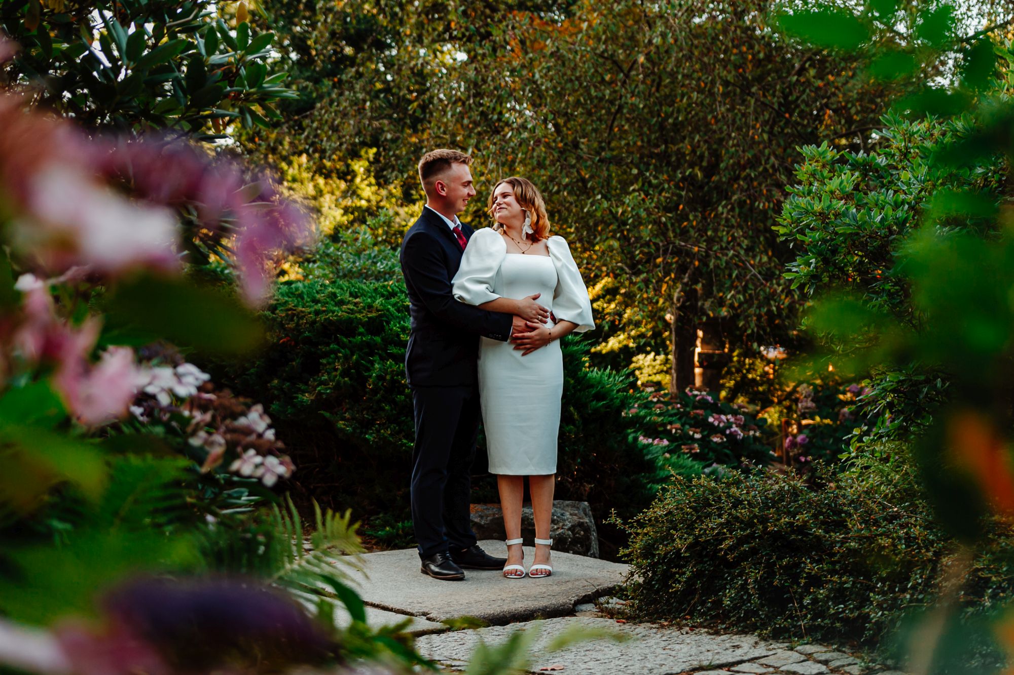 Sesja plenerowa Legnica – para młoda pozuje w ogrodzie, elegancki pan młody w garniturze obejmuje pannę młodą w białej sukni, otoczeni zielenią i kwiatami.